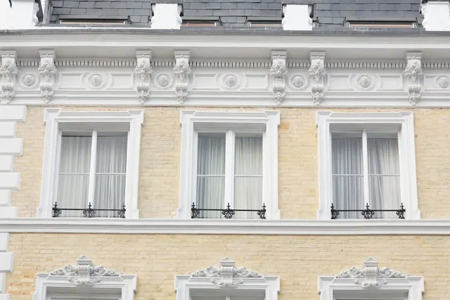 Exterior of a cream brick building with white ornate cornices, sash windows with sheer curtains and wrought iron window railings, below a slate roof.