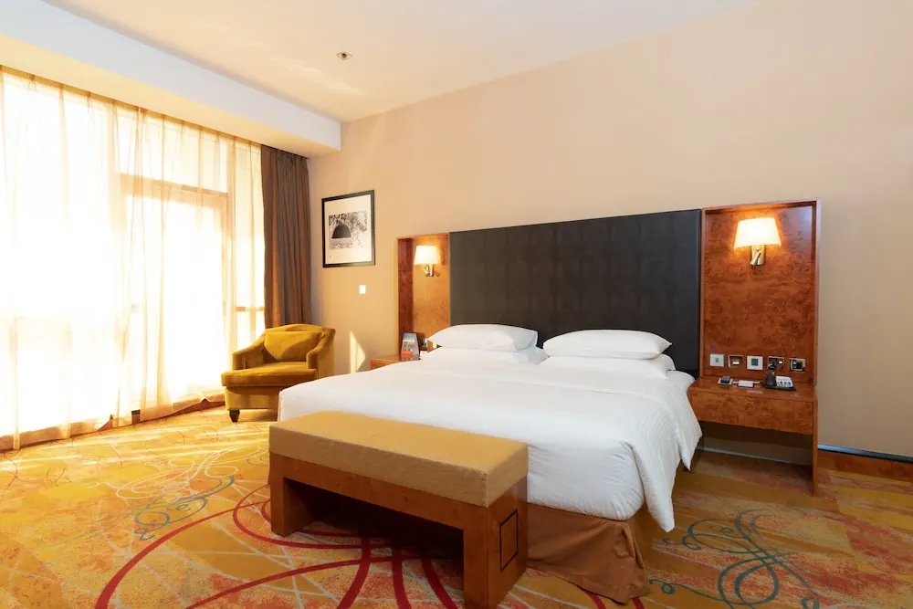 A king bed with a dark upholstered headboard, a wooden bench at its foot, a yellow armchair and sheer curtains covering a large window in a hotel bedroom.
