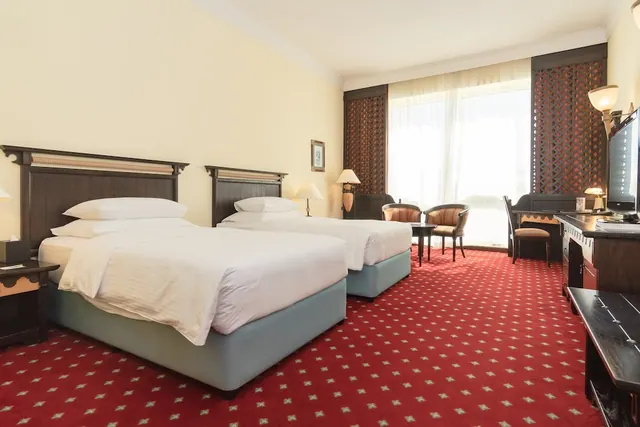 A hotel bedroom with two beds, armchairs, a work desk and a television, with a large window behind sheer curtains.