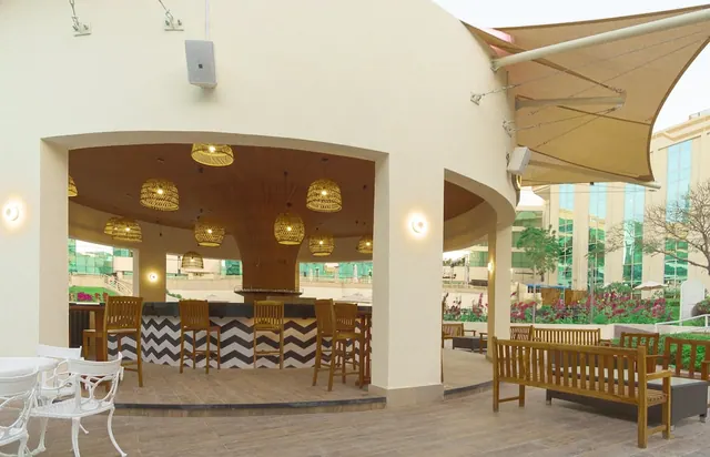 An outdoor bar area with wicker pendant lights, a chevron-patterned bar front, wooden benches and white metal chairs, with a garden and glass building visible beyond.