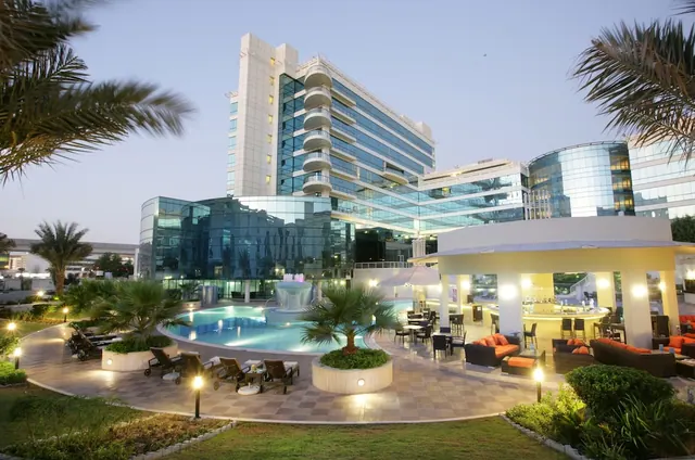 Outdoor pool area at dusk with sun loungers, a fountain, lounge seating and a pool bar in front of a glass-fronted hotel building.