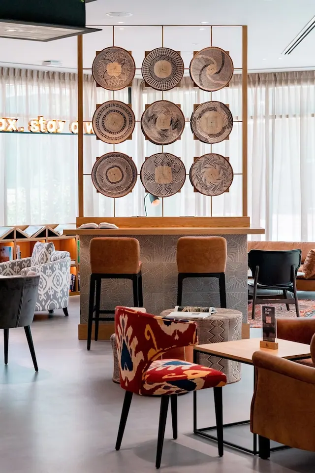 A lounge area with a wooden bar counter, two stools, woven baskets mounted on a divider, and colourful upholstered armchairs.