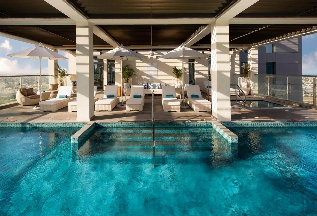 A large rooftop swimming pool with white sun loungers and shaded umbrellas under a covered terrace, with a small pool to the right and hazy city buildings beyond the railing.