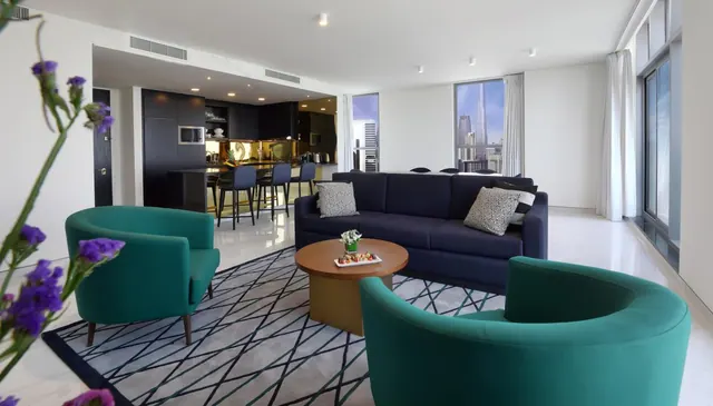 A living area with a navy sofa, two teal armchairs and a round wooden coffee table, with an open kitchen to the left and a window showing the Burj Khalifa tower.