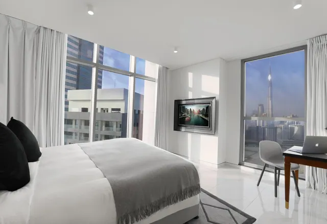A bedroom with a made king bed, a wall-mounted television, a work desk with a laptop, and floor-to-ceiling windows showing the Burj Khalifa and city towers.