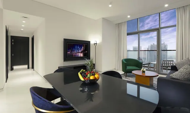 A living and dining area with a dark dining table holding a fruit bowl, a wall-mounted television, a green armchair, and floor-to-ceiling windows opening onto a balcony with a view of city towers.