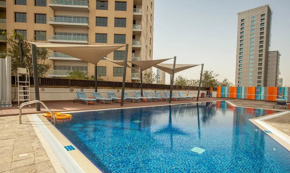 Outdoor swimming pool with blue tiles, striped sun loungers under shade sails, and high-rise buildings behind.