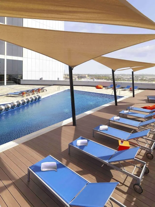 A rooftop swimming pool with blue sun loungers and white towels under beige shade sails, with a hazy low-rise city visible beyond the terrace.