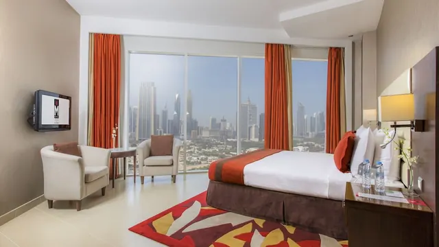 A bedroom with a double bed, two armchairs, a wall-mounted television and floor-to-ceiling windows overlooking a city skyline.