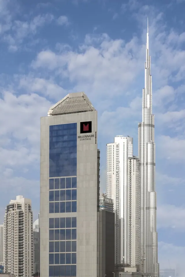 The Millennium Central hotel tower stands beside the Burj Khalifa under a partly cloudy blue sky.