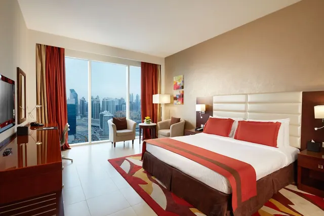 A hotel bedroom with a king bed, two armchairs, a wooden desk with a television, and floor-to-ceiling windows overlooking a city skyline.