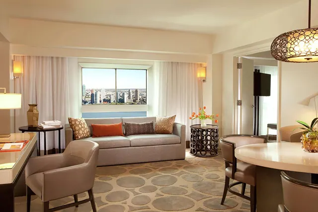 A staged hotel living area with a grey sofa with patterned cushions, a round dining table with chairs, and a window showing a city skyline.