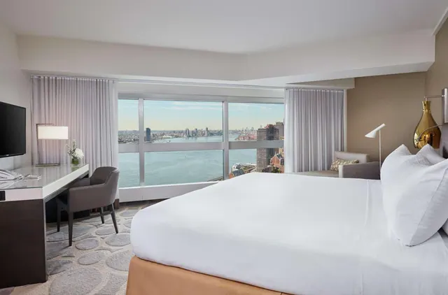 A king bed faces floor-to-ceiling windows with a view of the river and city skyline, beside a work desk and television.