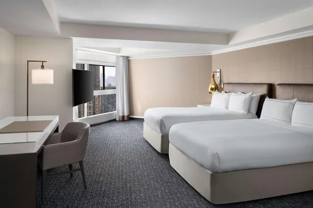 A bedroom with two queen beds, a desk and chair, a wall-mounted television, and a window showing tall city buildings.