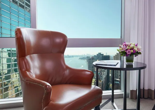 A brown leather armchair and a small side table with a flower vase sit in front of a window overlooking a river and city buildings.