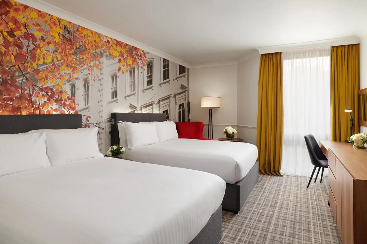 A hotel bedroom with two double beds, an autumn tree mural on the wall behind them, a red armchair, yellow curtains and a window with sheer drapes.