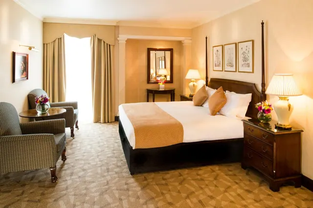 A bedroom with a king bed, two armchairs beside a small round table, bedside lamps, framed prints and tall curtained windows letting in daylight.