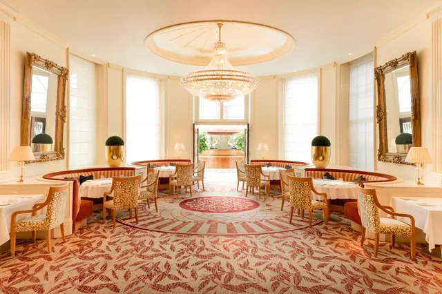 A dining room with curved banquette seating, tables laid with white linen, a large chandelier and ornate gilt mirrors, with an open doorway onto a courtyard with plants.
