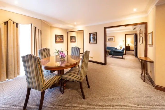 A round wooden dining table with striped upholstered chairs and a flower vase stands near a doorway leading to a seating area with blue armchairs and a sofa.