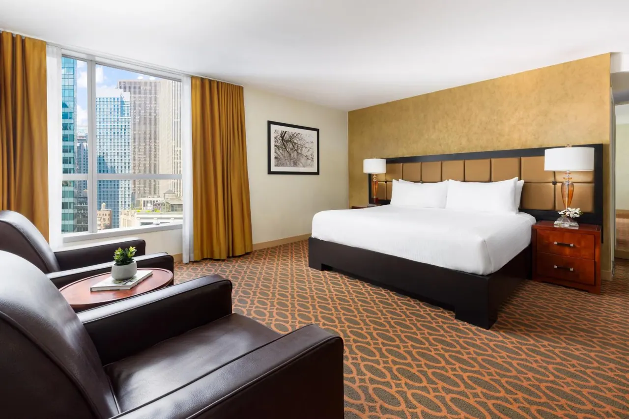 A hotel bedroom with a king bed, two leather armchairs by a window, and city skyscrapers visible outside.