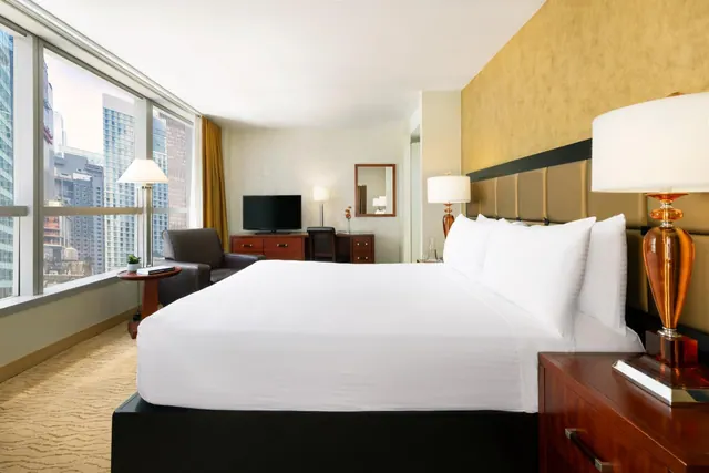 A hotel bedroom with a king bed, an armchair, a wooden desk and dresser with a television, and floor-to-ceiling windows looking out over city high-rises.