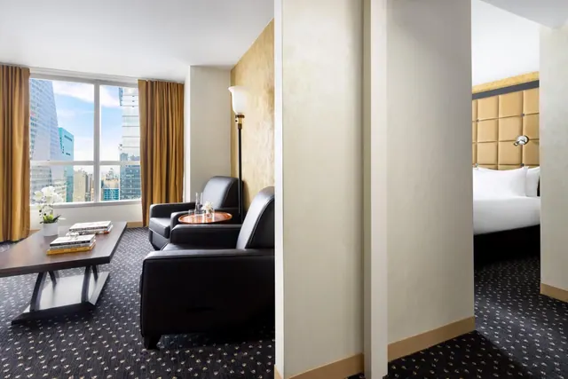 A living area with two leather armchairs, a small round side table and a dark coffee table, with a floor-to-ceiling window showing city buildings and an open doorway to a bed with a tufted headboard.