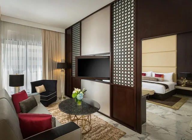 A living area with a grey sofa, an armchair, a round coffee table with flowers and a wall-mounted television, with a bed visible through the doorway in the background.