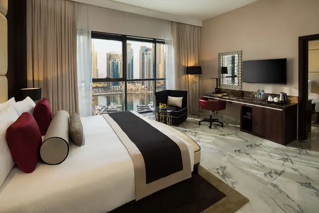 A bedroom with a king bed facing a window that looks out over marina towers and water, with an armchair, work desk and wall-mounted television on the right.