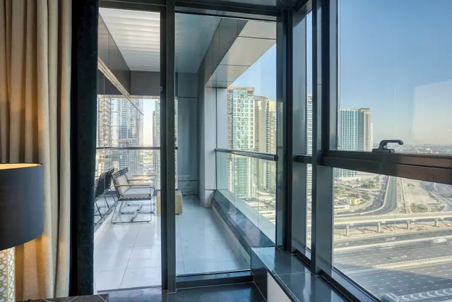 Glass sliding doors open onto a balcony with metal chairs, with high-rise towers and a multi-lane road visible through the adjacent window.