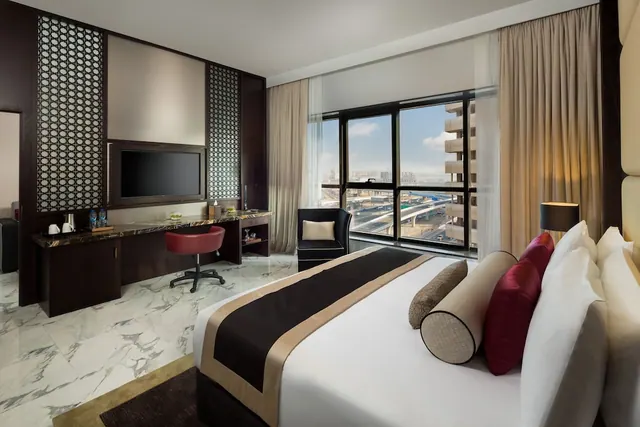 A hotel bedroom with a king bed, a desk with a red chair and wall-mounted TV, and floor-to-ceiling windows overlooking a highway and distant city buildings.