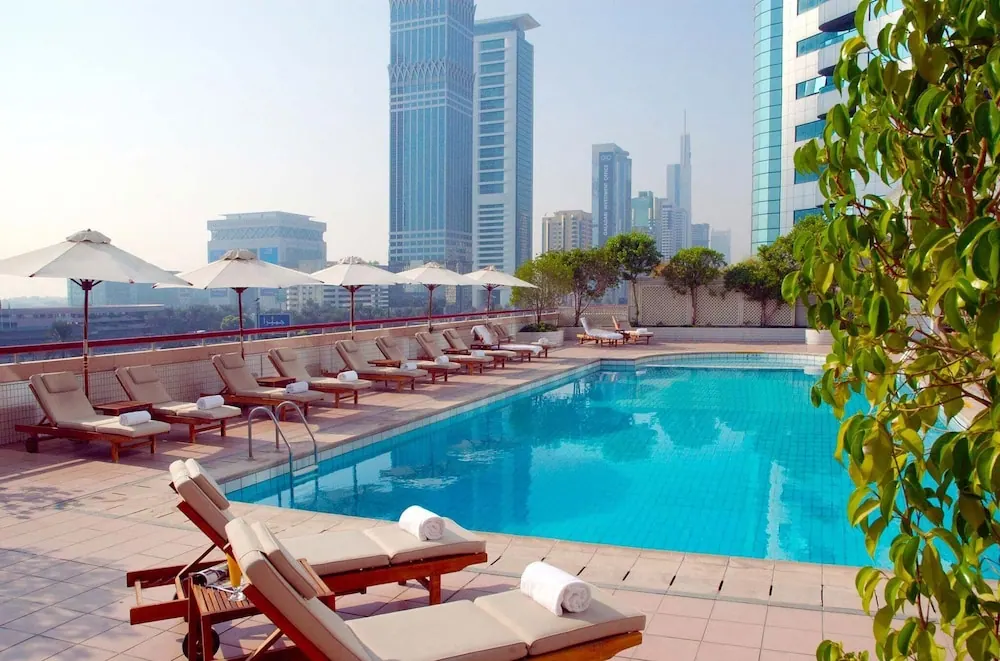 A rooftop swimming pool with white sun loungers and umbrellas, with city skyscrapers visible in the background.