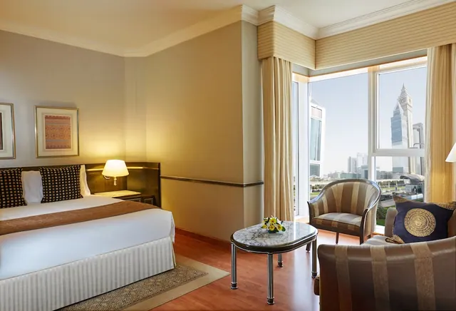 A hotel bedroom with a made-up double bed, an armchair seating area with a round table, and a large window looking out over a city skyline with a clock tower.