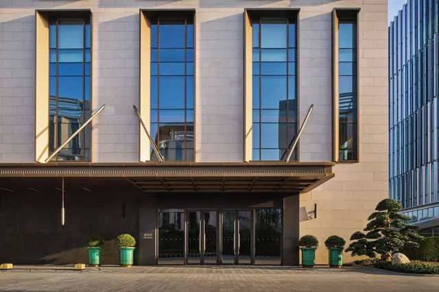 The entrance of a stone-clad building with tall windows, glass double doors, green planters and a trimmed tree on a paved forecourt.