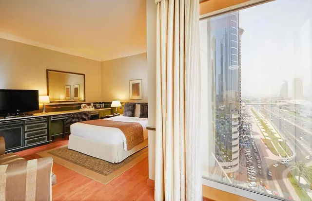 A hotel bedroom with a double bed, a desk and a television, and a large window overlooking a busy road with tall towers beyond.