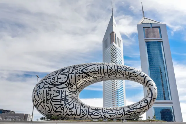 The Museum of the Future, a torus-shaped building covered in Arabic calligraphy, stands in front of the Emirates Towers under a partly cloudy sky.