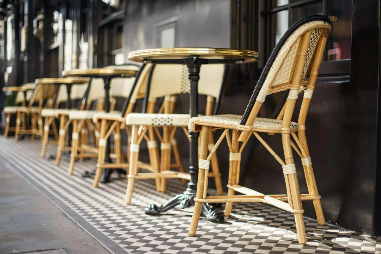 Empty rattan bistro chairs and small round brass-topped tables stand on a checkered tile floor along an outdoor terrace.