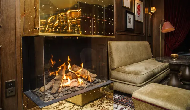 A lounge area with a glass-fronted gas fireplace, a green velvet sofa, a round marble side table and a pink-shaded floor lamp.