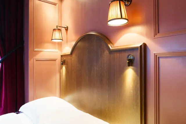 A bedroom with a wooden arched headboard, two pleated wall lamps on pink panelled walls, a burgundy curtain at the left edge, and white bedding.