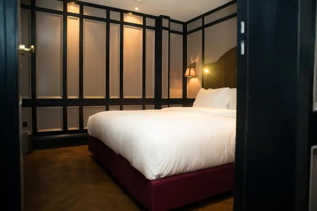 A double bed with white linen and a dark red base stands against dark panelled walls in a bedroom, photographed through an open door.