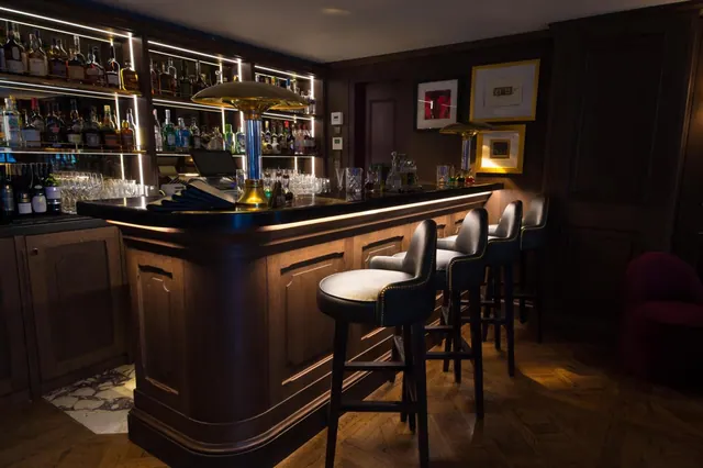 A dimly lit bar with a curved wooden counter, leather-upholstered bar stools, and shelves of spirit bottles backlit by strip lights.