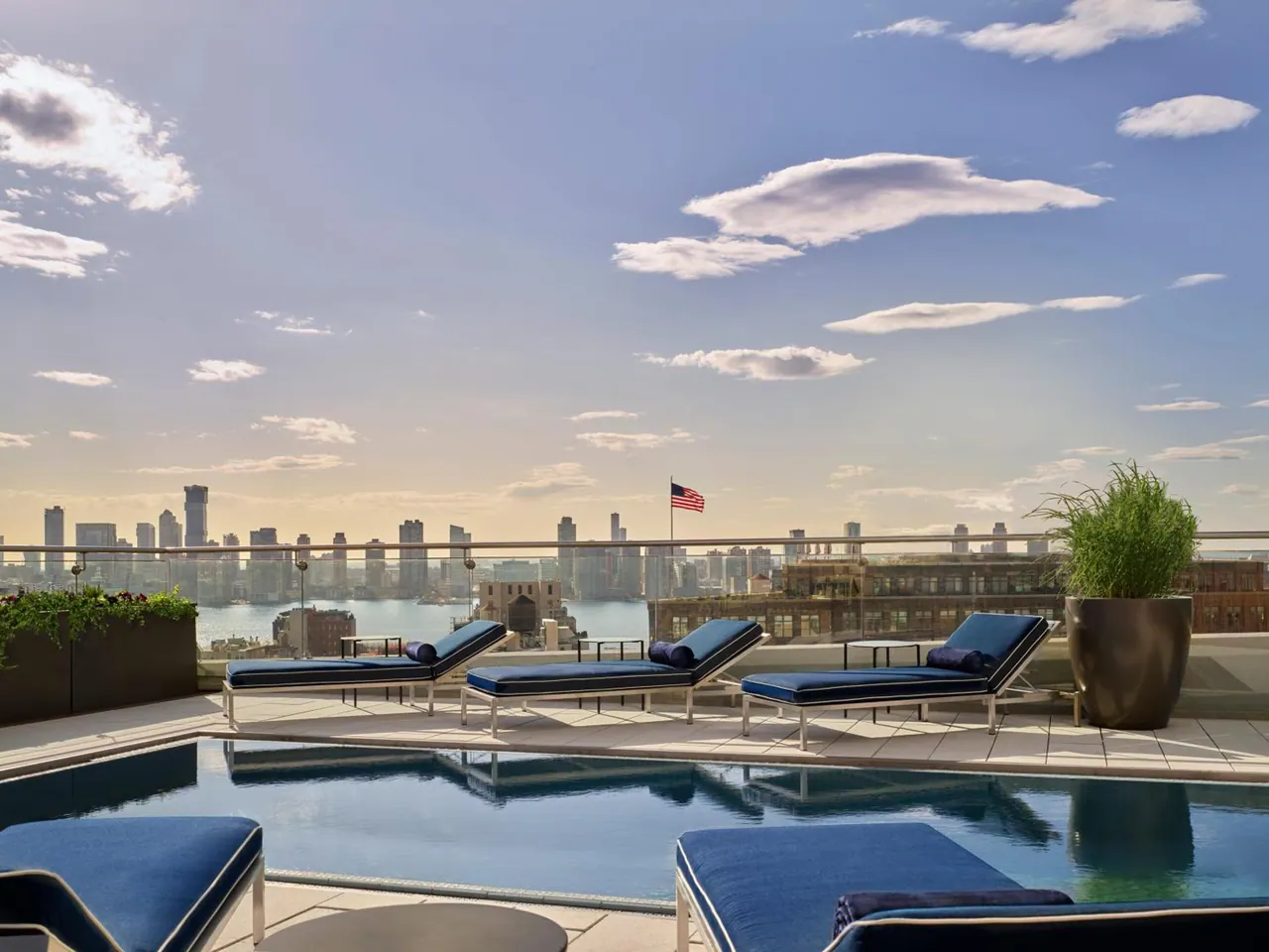 A rooftop pool with navy loungers overlooks a city skyline and a river under a partly cloudy sky.