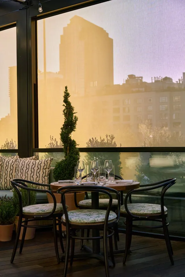 A set round dining table with wine glasses and cushioned chairs sits on a glass-enclosed terrace, with city buildings visible through the windows.
