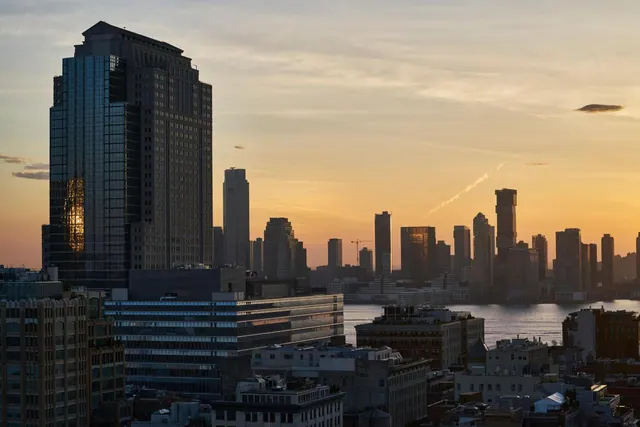 A city skyline with tall buildings and a river is shown at sunset, viewed from above.