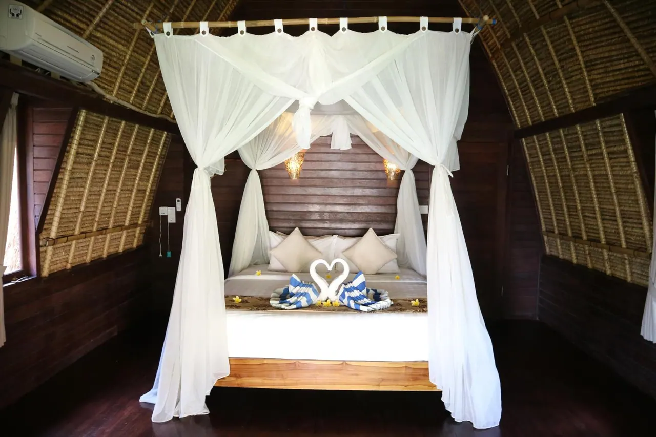 A canopy bed with white mosquito netting and folded towel swans sits in a room with a thatched bamboo ceiling and a wall-mounted air conditioner.