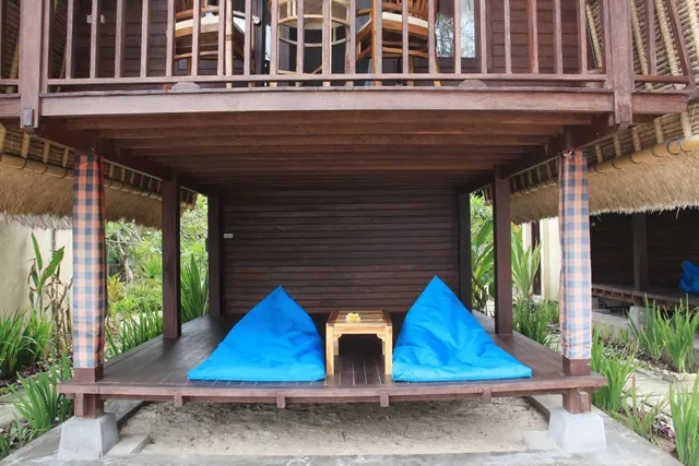 A raised wooden terrace under an upper balcony has two blue bean bag cushions and a small wooden table beside a thatched-roof bungalow.
