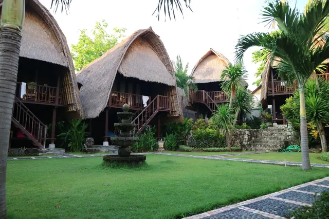 Thatched-roof bungalows with wooden balconies and staircases stand around a trimmed lawn with a stone fountain and palm trees.