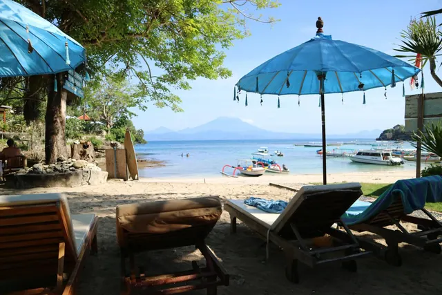 Sun loungers and blue umbrellas sit on a sandy beach facing the sea, with moored boats and a mountain in the distance.