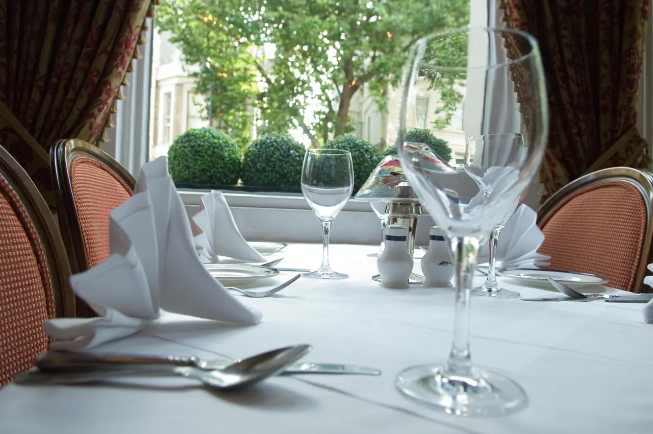 A dining table laid with white linen, wine glasses, folded napkins and cutlery, with a window behind showing trees and topiary bushes outside.