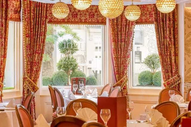 A restaurant dining room with red patterned drapes, crystal chandeliers, and set tables with folded napkins and wine glasses, with windows onto a street with topiary planters.