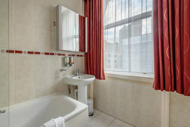 A bathroom with a white bath, pedestal sink, mirror cabinet, cream tiled walls and red curtains framing a window that looks out onto tall buildings.